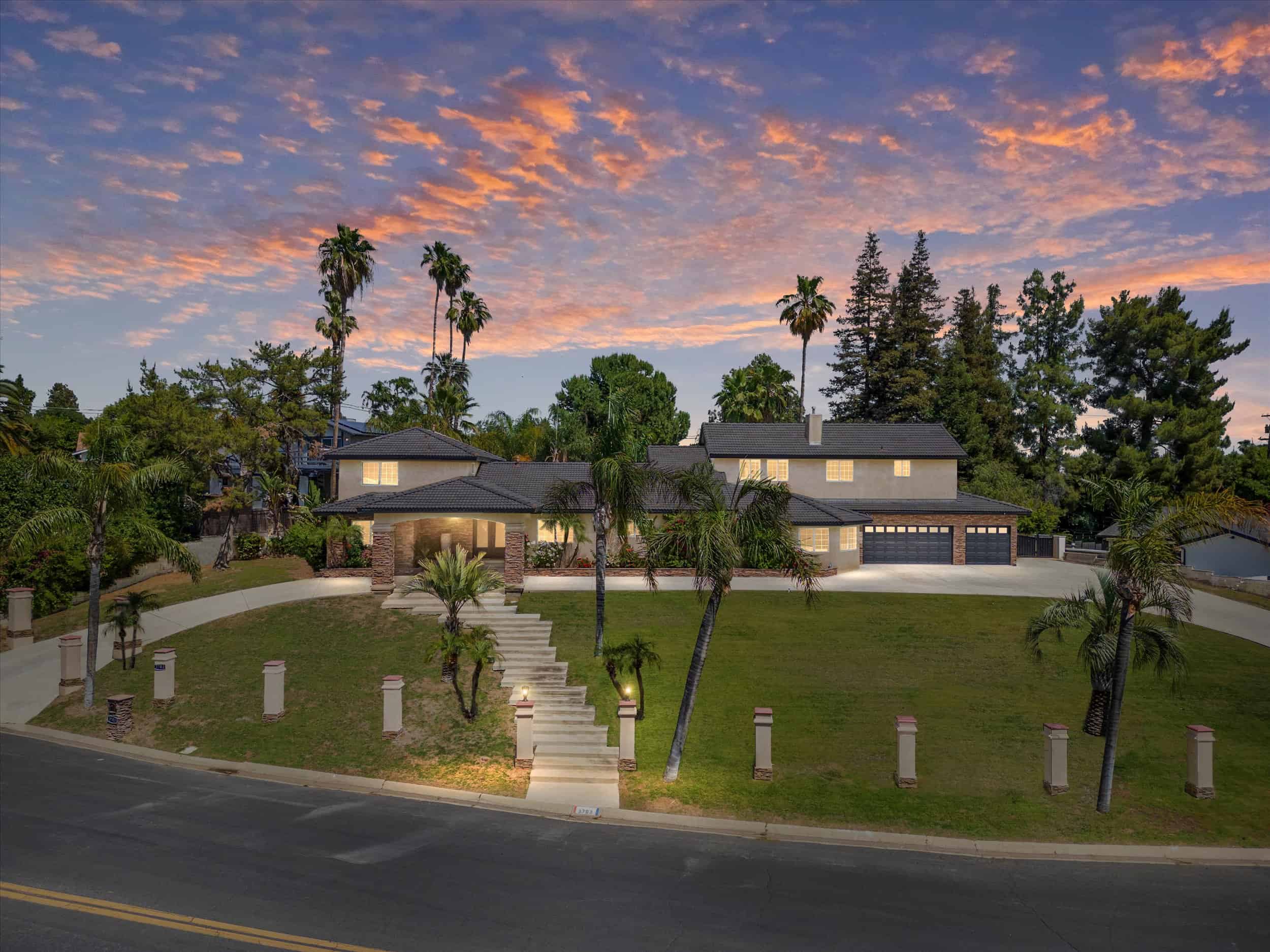 Home exterior at 3703 Braeburn Dr. showcases a large house, palm trees, stairs, and a vibrant sunset. Real estate photography.
