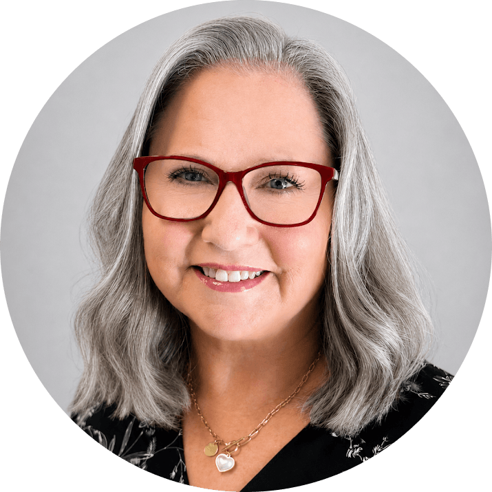 Catherine 1 - Bakersfield Recovery Center Smiling woman with shoulder-length gray hair and red glasses, wearing a black patterned top and heart pendant necklace.