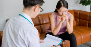 A clinician in a striped shirt with a stethoscope talks to a worried woman on a brown sofa, taking notes in a binder.