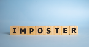 Wooden letter blocks spell 'IMPOSTER' on a light-blue background, likely illustrating impostor themes or impostor syndrome.