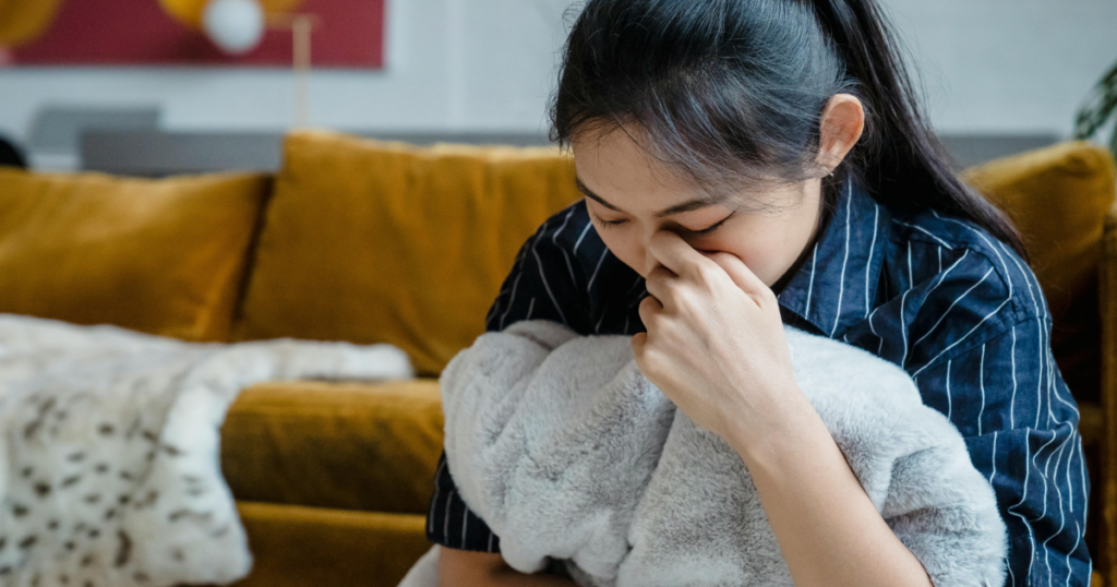 Woman seated on a brown couch, clutching a soft gray blanket and covering her face with her hand, appearing upset.