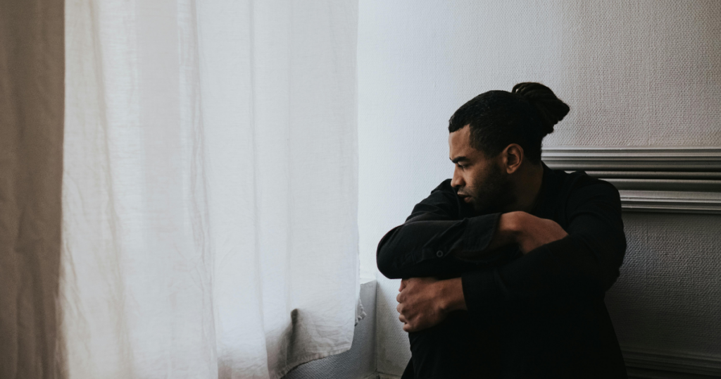 Man sits by a window with white curtains, arms wrapped around his knees, looking down in thought or sadness.