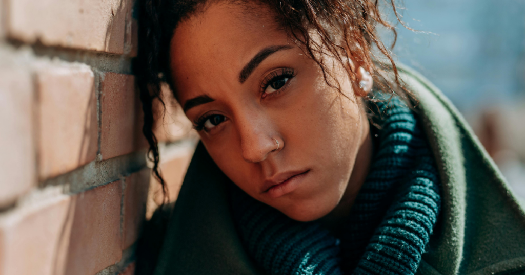 Woman with dark hair and a nose ring leans against a brick wall, wearing a teal scarf and green coat, gazing at the camera.