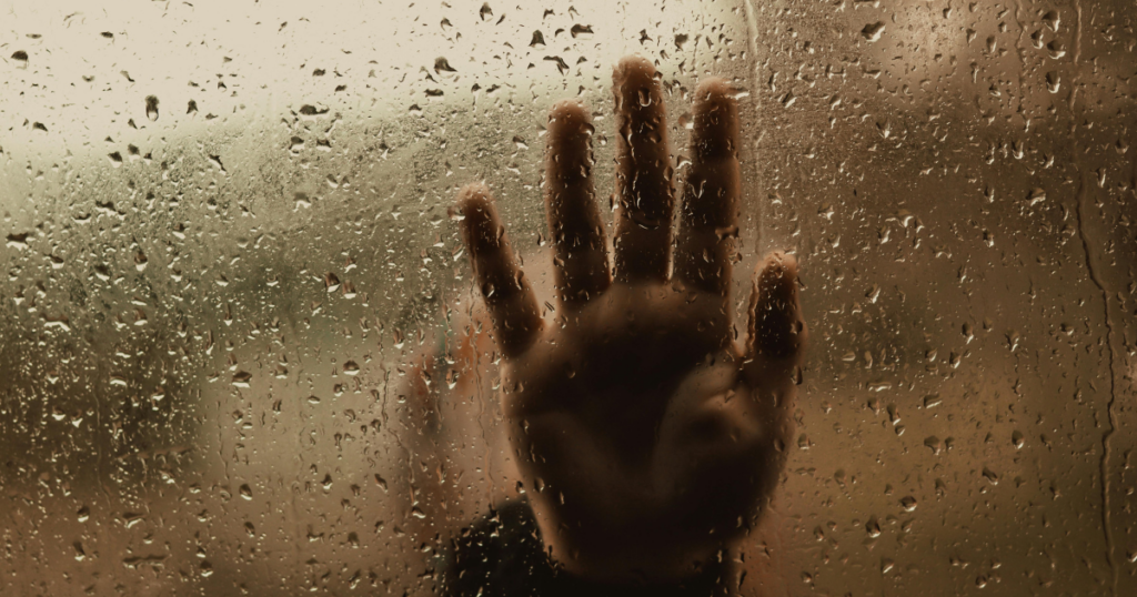 Hand pressed against a rain-speckled glass window, suggesting separation or longing.