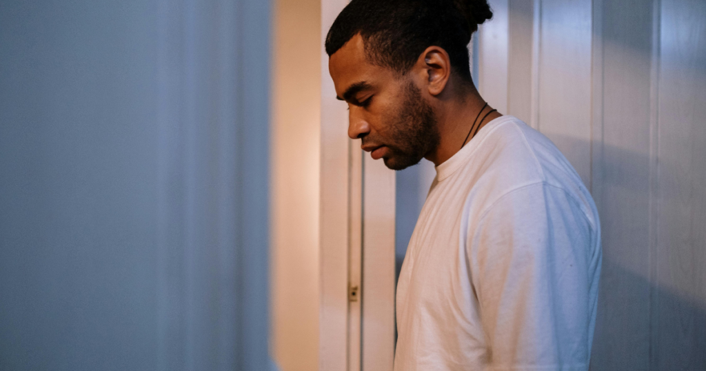 Young man in a white T-shirt leaning against a wall in a dim hallway, looking down in thought.