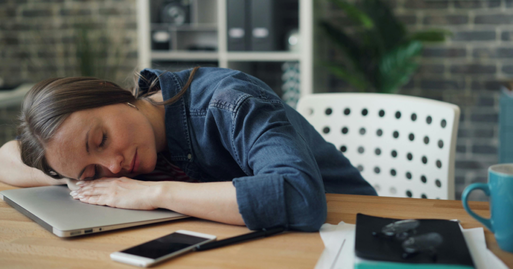 Woman asleep with her head on a laptop on a wooden desk, with a phone, notebook, pen, and blue mug nearby.