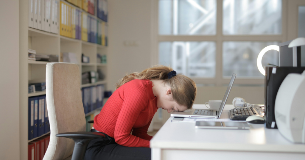 Person with a red sweater rests head on a laptop at a bright office desk, surrounded by shelves and files.