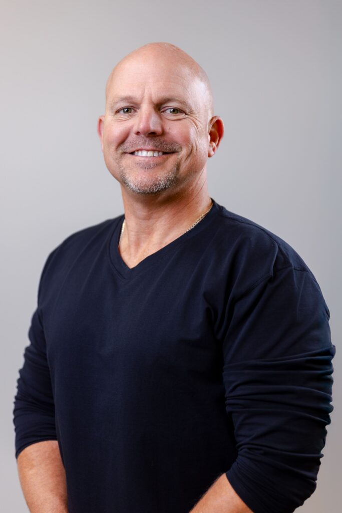 Smiling bald man in a dark crew-neck shirt against a neutral gray background (portrait).