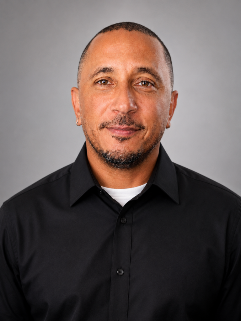 Close-up portrait of a smiling man with short hair and a goatee, wearing a black button-down shirt against a gray backdrop.